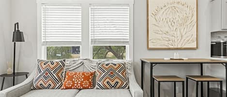 Una sala moderna con un sofá gris, cojines estampados, una mesita auxiliar, una lámpara de pie y una zona de cocina con mostrador y armarios.