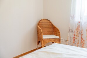 Een rieten stoel met een kussen tegen een muur, een bed met een wit laken, en een stopcontact in de muur.