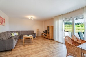 A modern living room with a grey sofa, wooden coffee table, and a wicker chair. A wooden cabinet and a television stand are also visible. The room has large windows offering a view of a green landscape. - Apartment 'Ferienwohnung Rebblüte' with Mountain View, Shared Garden and Wi-Fi (Oberkirch)