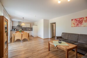 Een moderne woonkamer met houten vloer, een bank, een eettafel met stoelen en een keuken op de achtergrond.