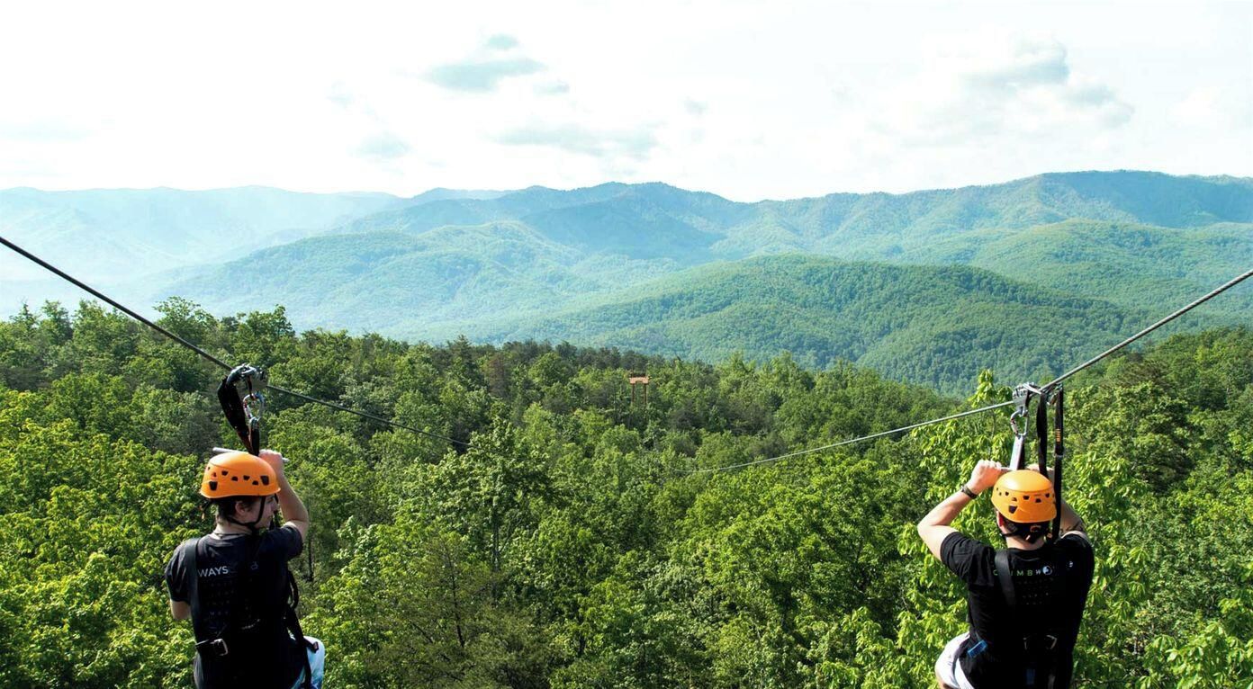 Zwei Personen auf einer Zipline vor einer bewaldeten Berglandschaft.