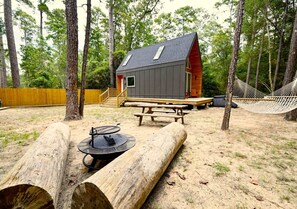 A cabin with a sloped roof, a wooden deck, a picnic table, a hammock, and a fire pit.