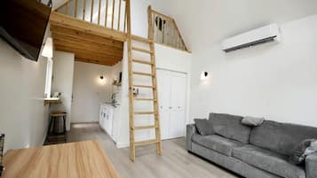 A loft-style room with a wooden staircase, a grey sofa, a wooden dining table, and a kitchen area in the background.
