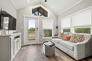 A modern living room with a grey sofa, a coffee table, a flat-screen TV, and a large window with blinds. - Pet Friendly Cabin with Fire Pit in Fredericksburg, Texas (Fredericksburg)