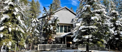 Un chalet enneigé doté d’une terrasse en bois et d’une grande fenêtre donnant sur un paysage hivernal.