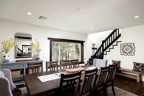 A dining area with a dark wooden table, chairs, and a large window. A staircase with a black railing is visible in the background. - Luxurious Cabin with Game Room, Amazing Deck and Spa in California (Big Bear Lake)