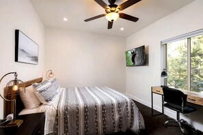 Une chambre à coucher comprenant un lit, un bureau, une chaise, un ventilateur de plafond et une fenêtre donnant sur des arbres.