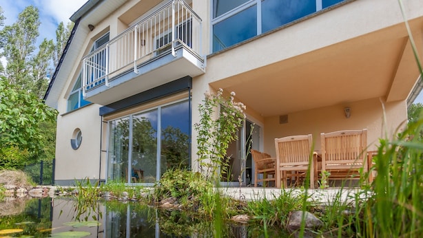 A modern building with a glass facade, a balcony with a railing, and a small pond with plants and rocks. - Modern vacation home with plenty of space to relax - Haus Eleganza (Glowe)