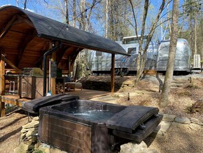 A wooden cabin with a covered patio, a hot tub, and a large RV in the background. - Luxury Airstream Rental for Intimate Getaways to Pisgah Forest, North Carolina (Pisgah Forest)