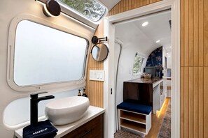 A compact bathroom with a sink, mirror, and a small seating area. - Luxury Airstream Rental for Intimate Getaways to Pisgah Forest, North Carolina (Pisgah Forest)