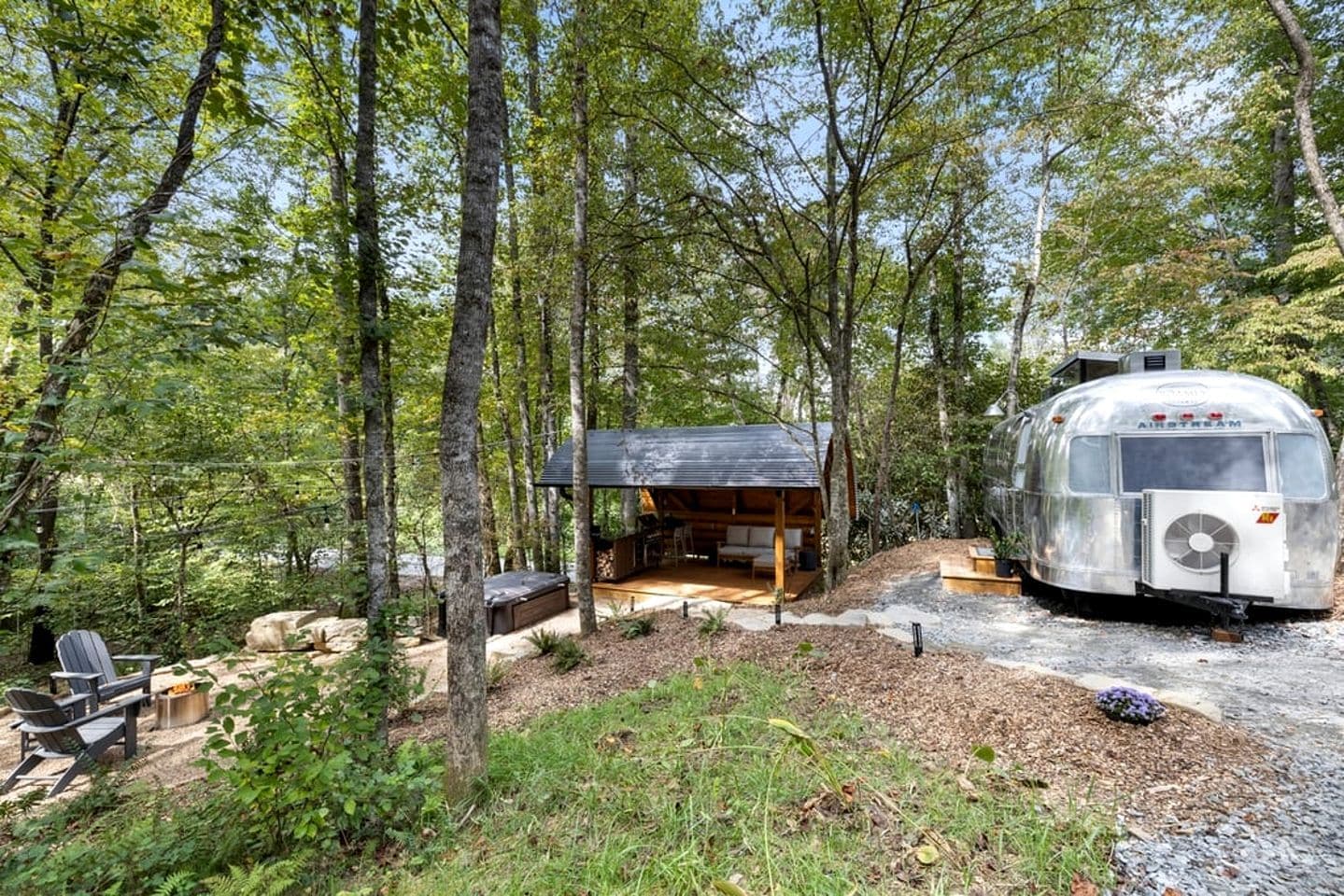 Eine rustikale Holzhütte mit überdachter Veranda und einem Oldtimer-Wohnwagen in bewaldeter Landschaft.