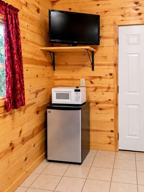 Habitación compacta con un televisor montado en una pared de madera, un microondas sobre un refrigerador pequeño y una puerta blanca.