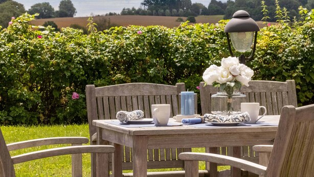 Una mesa de madera con sillas dispuestas al aire libre, una lámpara y un florero con flores blancas.