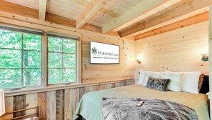 Camera da letto in una baita di legno con un letto, cuscini, una TV a parete e una finestra vista alberi.
