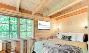 Une chambre à coucher dans une cabane en bois, avec un lit, des oreillers, un téléviseur fixé au mur et une fenêtre donnant sur la forêt.