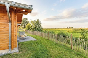 Ein Holzhaus mit überdachter Veranda, umzäunter Garten und Blick auf ein grünes Feld.