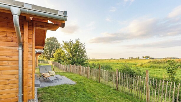 Ein Holzhaus mit überdachter Veranda, umzäunter Garten und Blick auf ein grünes Feld.