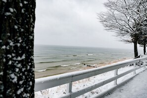 A snowy beach with a white railing, ocean waves, and bare trees.