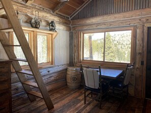 A rustic cabin interior with a wooden staircase, a table with chairs, a barrel, and a window with a forest view.