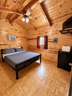 Habitació d'una cabana de fusta amb un llit, un ventilador de sostre, un calefactor i una finestra amb cortines.
