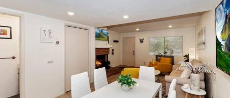 A modern living room with a fireplace, a dining area with a white table and chairs, and a yellow armchair.