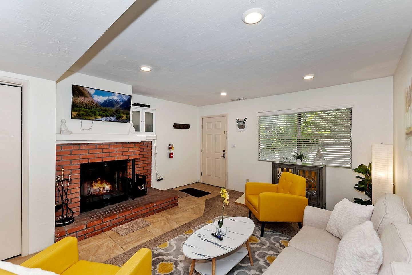 A living room with a fireplace, yellow armchairs, a white sofa, and a TV mounted above the fireplace.