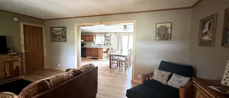 A living room with a brown sofa, a dark armchair, and a wooden cabinet. There is a kitchen visible through an open doorway.