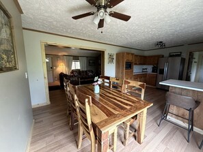 Un coin repas avec une table et des chaises en bois, un ventilateur de plafond et une cuisine en arrière-plan.