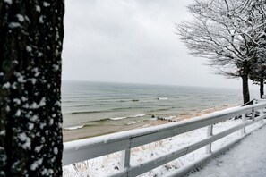 A snowy beach with a white railing, ocean waves, and bare trees.