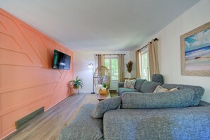 A living room with a sectional sofa, a flat-screen TV mounted on the wall, a large window with curtains, and a painting on the wall. - Lovely Beach House Rental in Coastal Michigan, for Memorable Getaways to Muskegon (Muskegon)