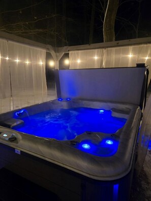 Un bain à remous avec un éclairage bleu, entouré d’une structure blanche, et un arbre en arrière-plan.