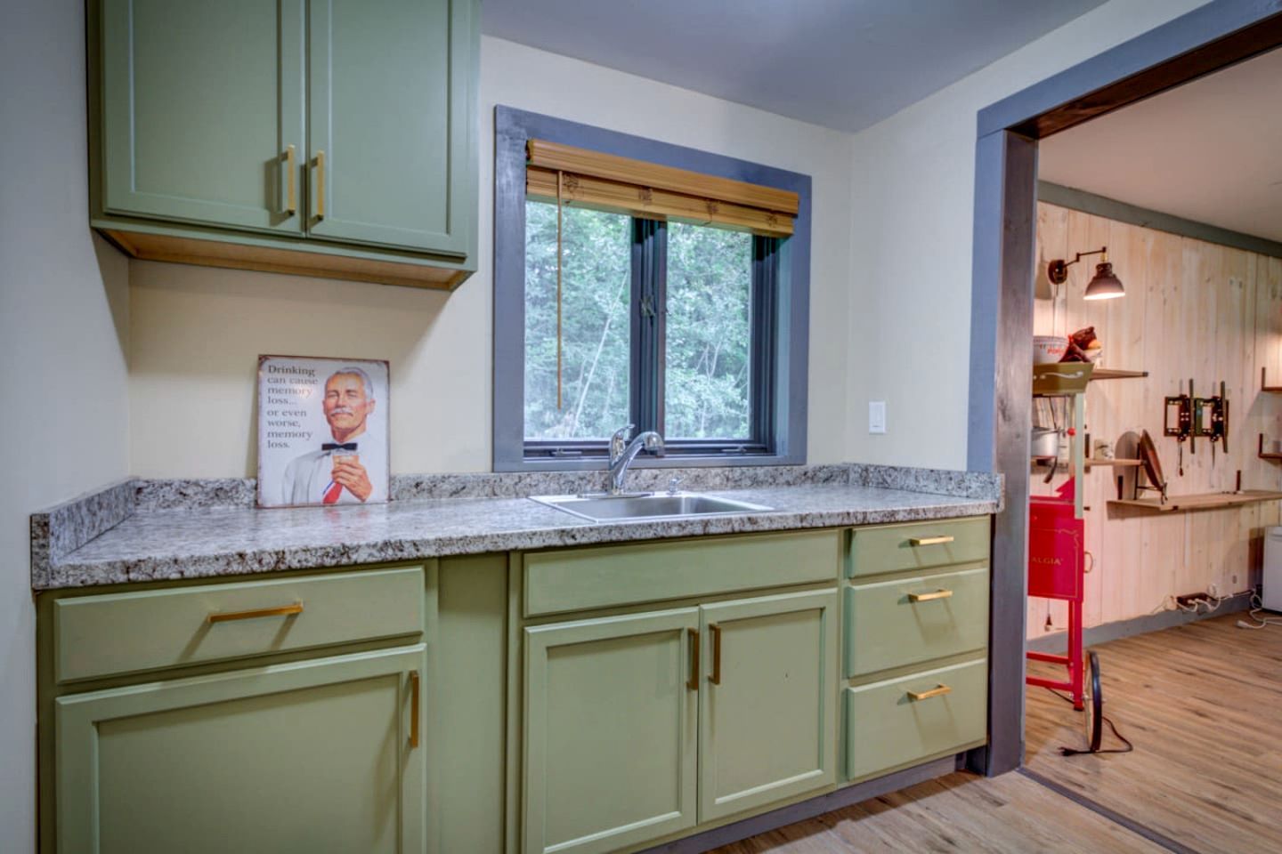 Una cocina con gabinetes verdes, encimera de granito, ventana con persiana de madera y un electrodoméstico rojo al fondo.
