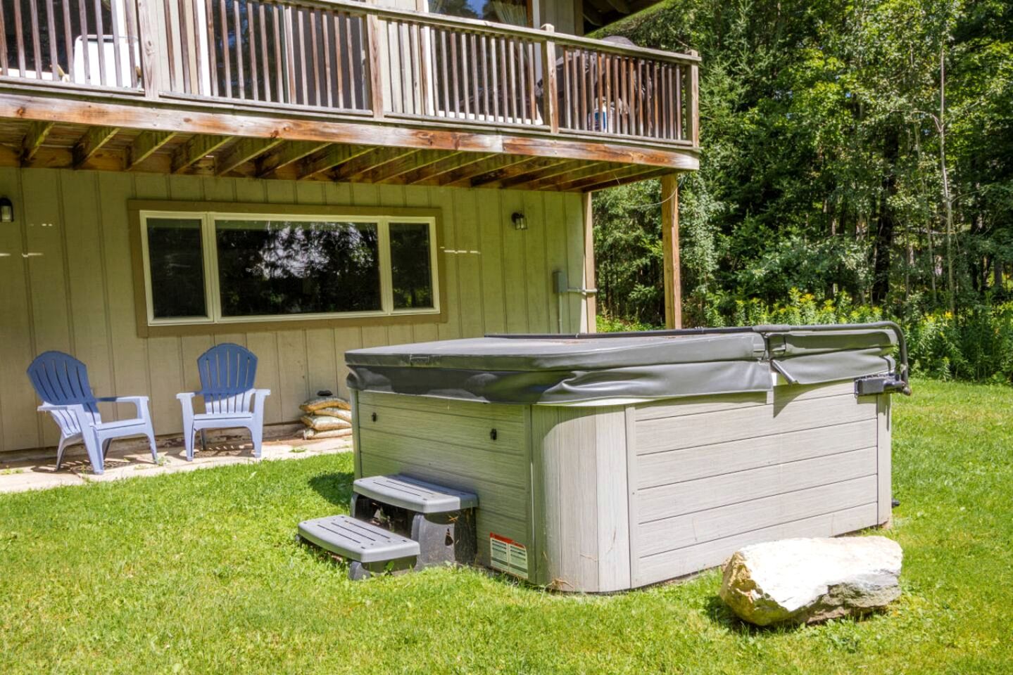 A hot tub in a backyard with a wooden deck, two Adirondack chairs, and a stone.