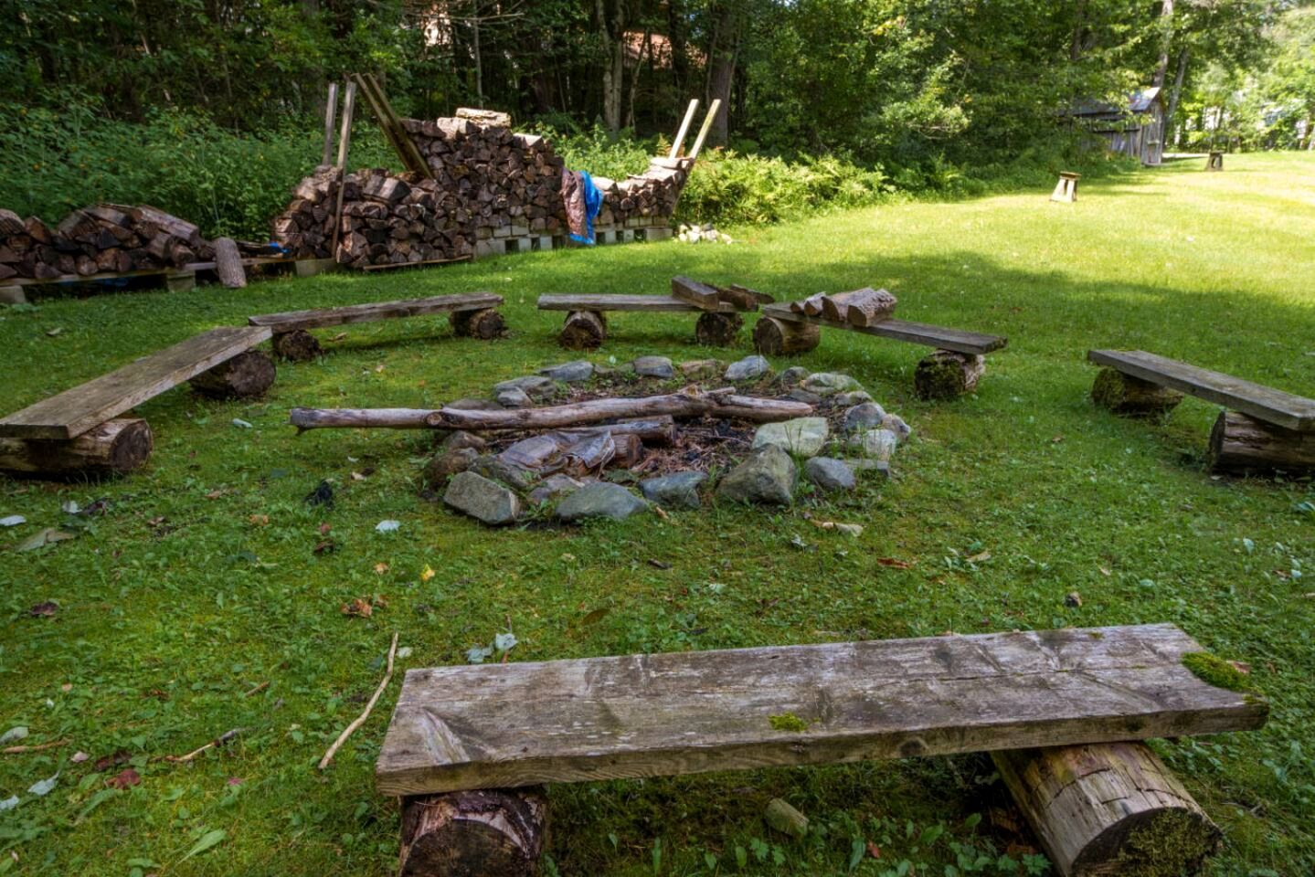 Disposición circular de troncos y piedras, bancos de madera y un montón de leña en un área verde al aire libre.