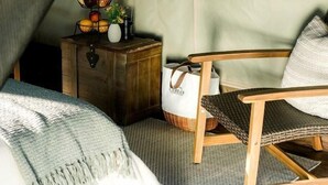 Een tent met een bed, een stoel, een klein houten kistje en een raam met uitzicht op bomen.