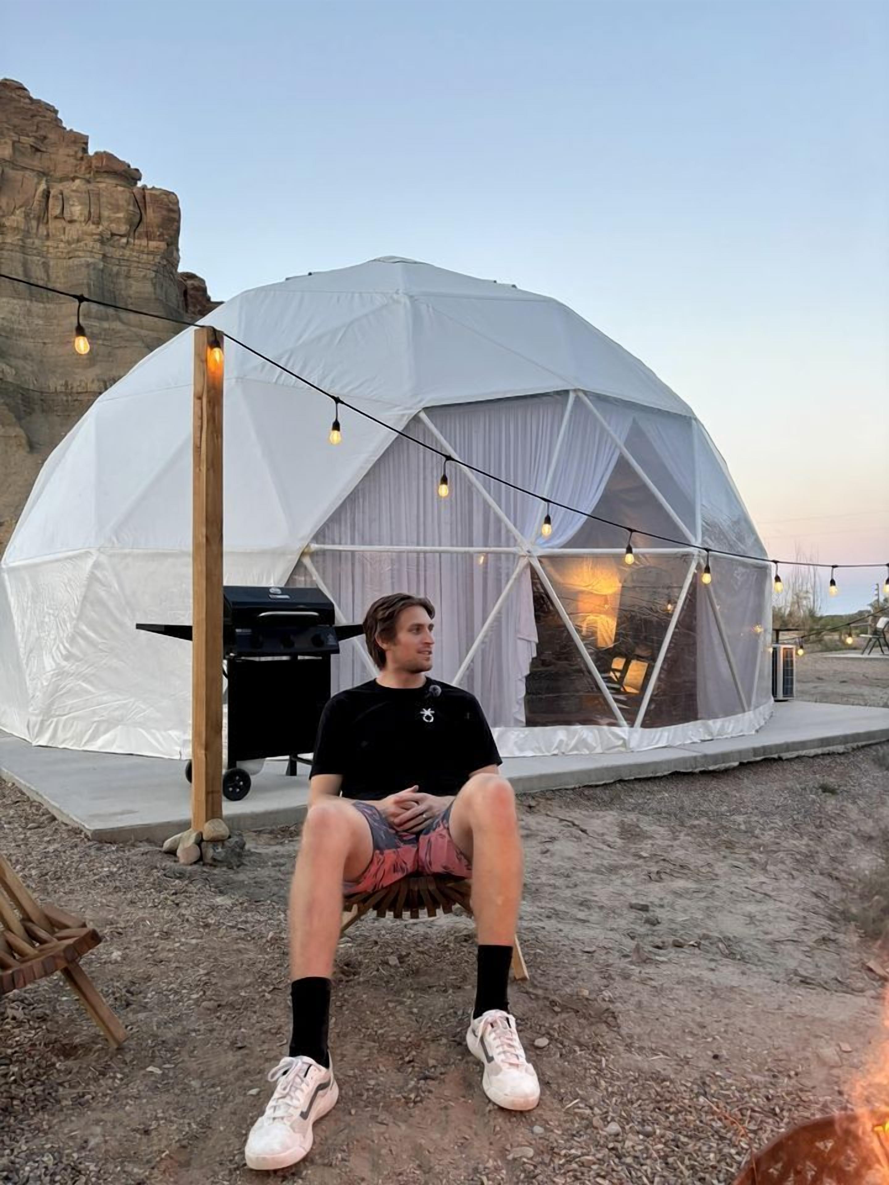 A person sitting outside a dome-shaped tent with a grill and string lights.