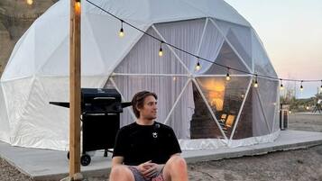 A person sitting outside a dome-shaped tent with a grill and string lights.