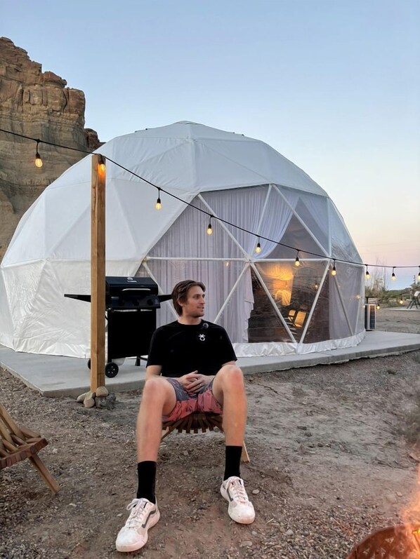A person sitting outside a dome-shaped tent with a grill and string lights.