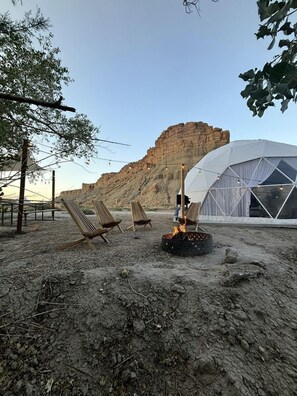 A desert landscape with a large white tent, wooden chairs, and a fire pit.