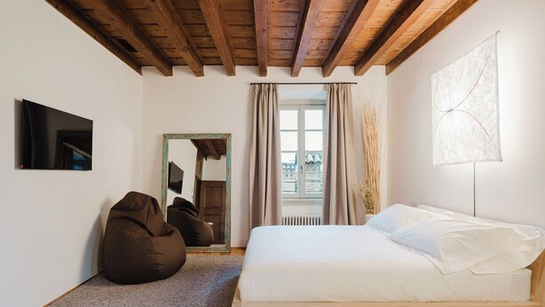 A bedroom with a wooden ceiling, a bed with white linens, a bean bag chair, a mirror, and a window with curtains. - Orto Room (Paladina)