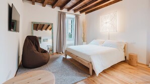A bedroom with a wooden ceiling, a bed, a bean bag chair, a small table, and a window with curtains. - Orto Room (Paladina)