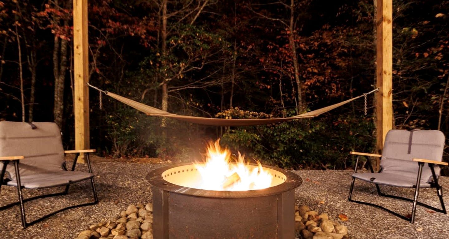 A fire pit with a hammock between two chairs in a wooded outdoor setting.