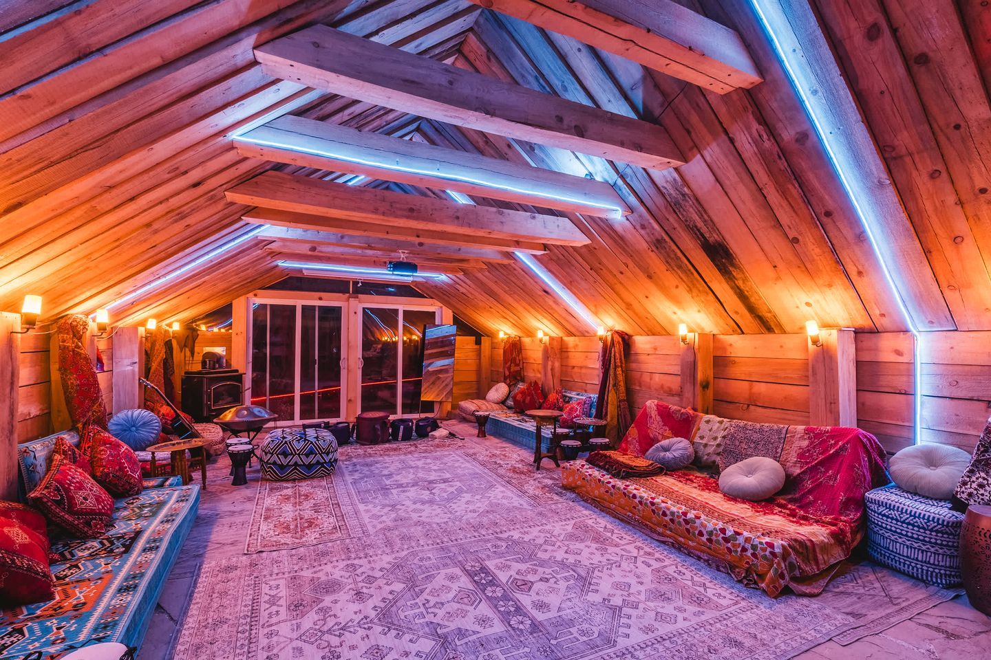 Ein Dachzimmer mit geneigter Decke, Holzbalken und einer zentralen Sitzecke mit Kissen und Teppichen.