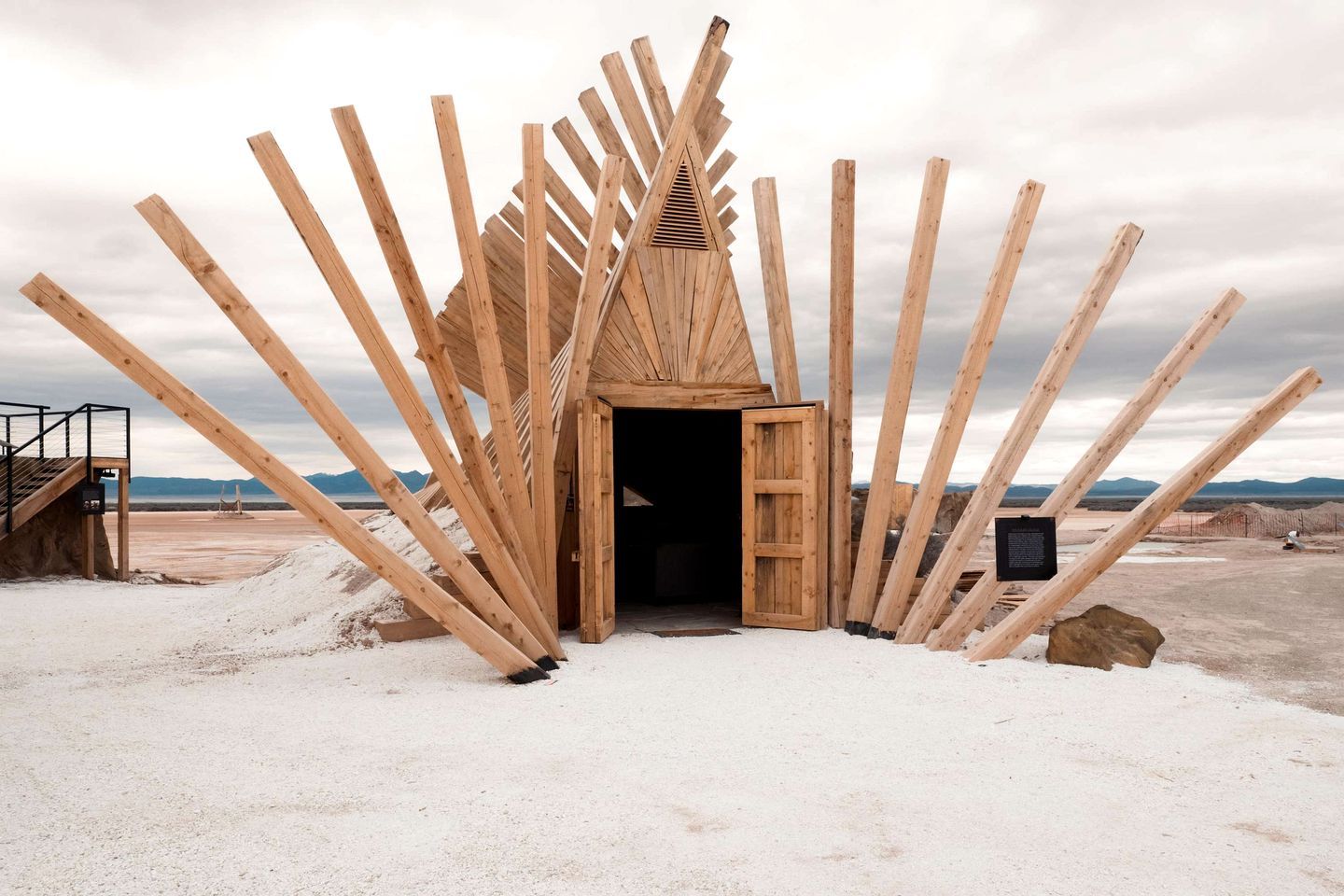 Estructura de madera con techo triangular y una puerta, rodeada de vigas de madera, ubicada en un paisaje árido con montañas al fondo.