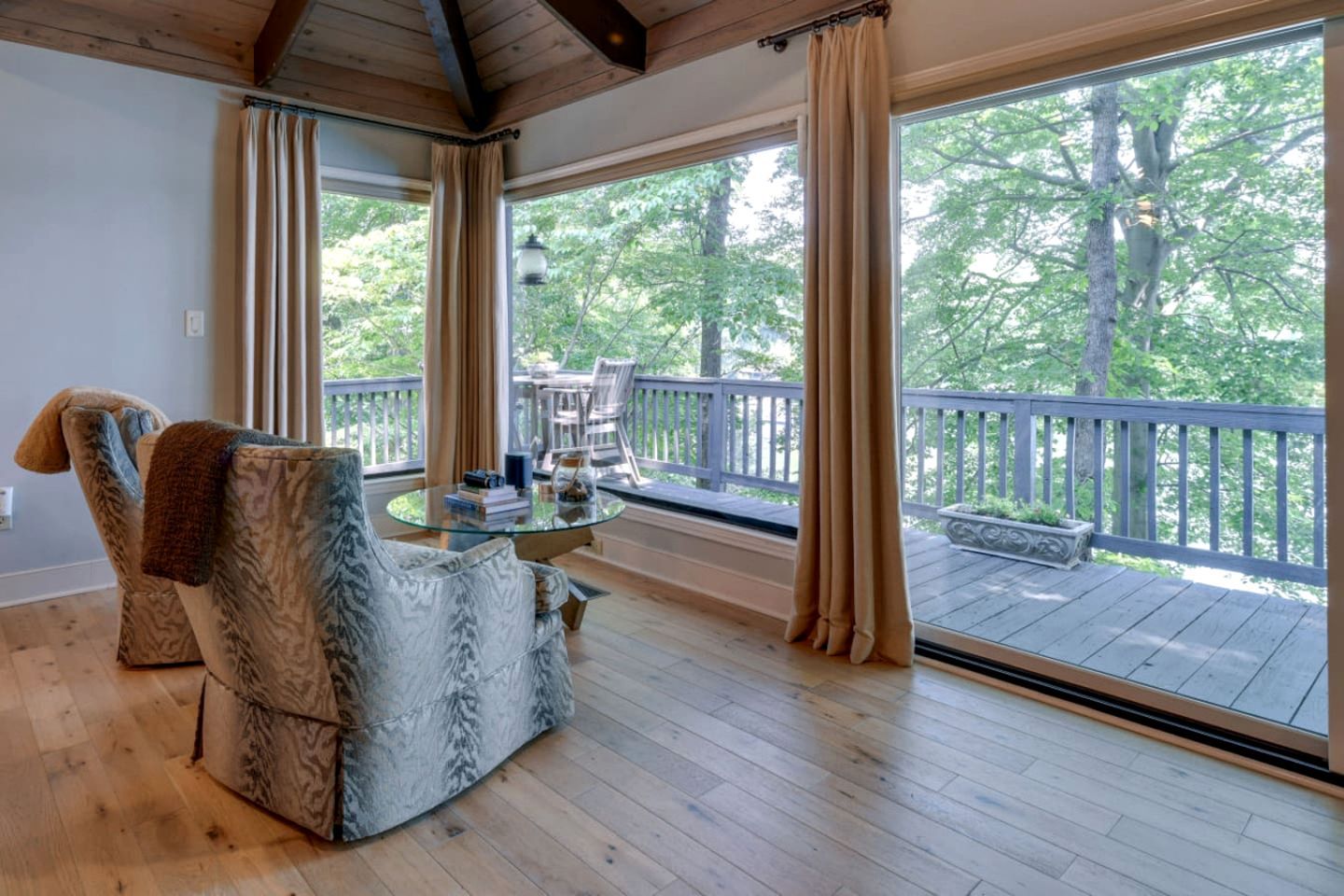 Ein Zimmer mit einem Glastisch, zwei gemusterten Sesseln und Blick auf eine Holzterrasse sowie Bäume durch große Fenster.