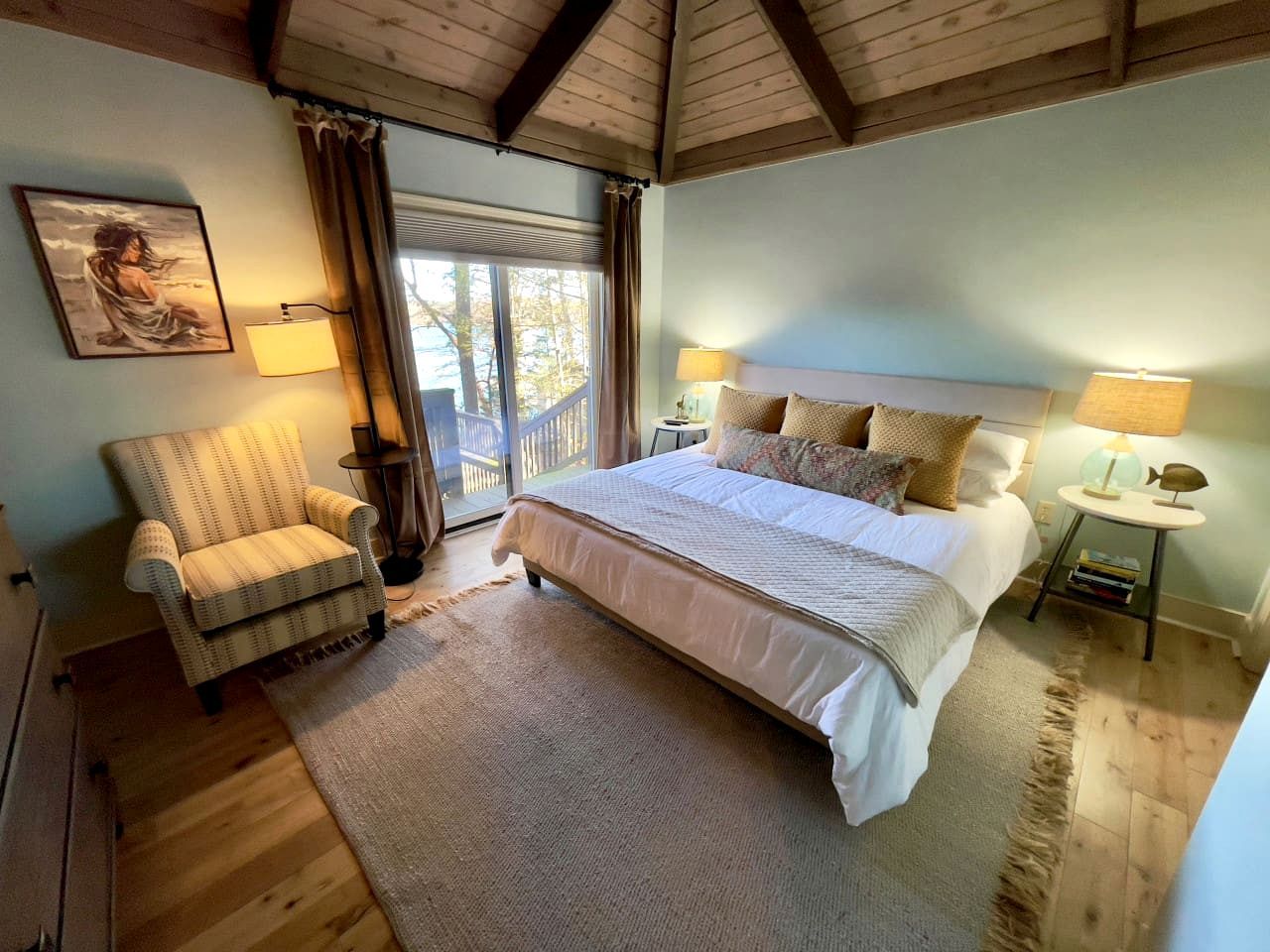 Quarto com cama, duas mesas de cabeceira, uma cadeira, janela com cortinas e um quadro na parede.