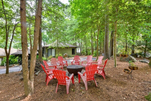 A forested area with a circular arrangement of red chairs around a central fire pit, surrounded by trees.