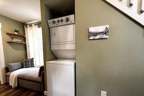 A compact living space with a bed, a small sofa, a washing machine, and a shelf with books. - Fully Stocked Cabin Nestled Among the Trees in Tennessee (Monteagle)