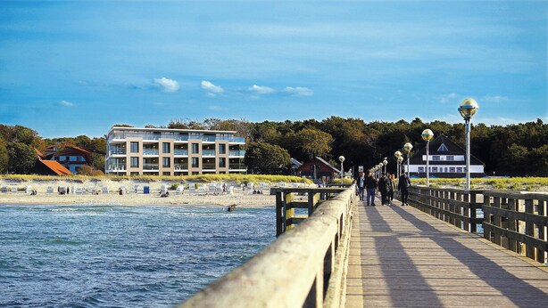 Ein hölzerner Steg führt zu einem Strand, im Hintergrund ein modernes Gebäude und ein Haus.