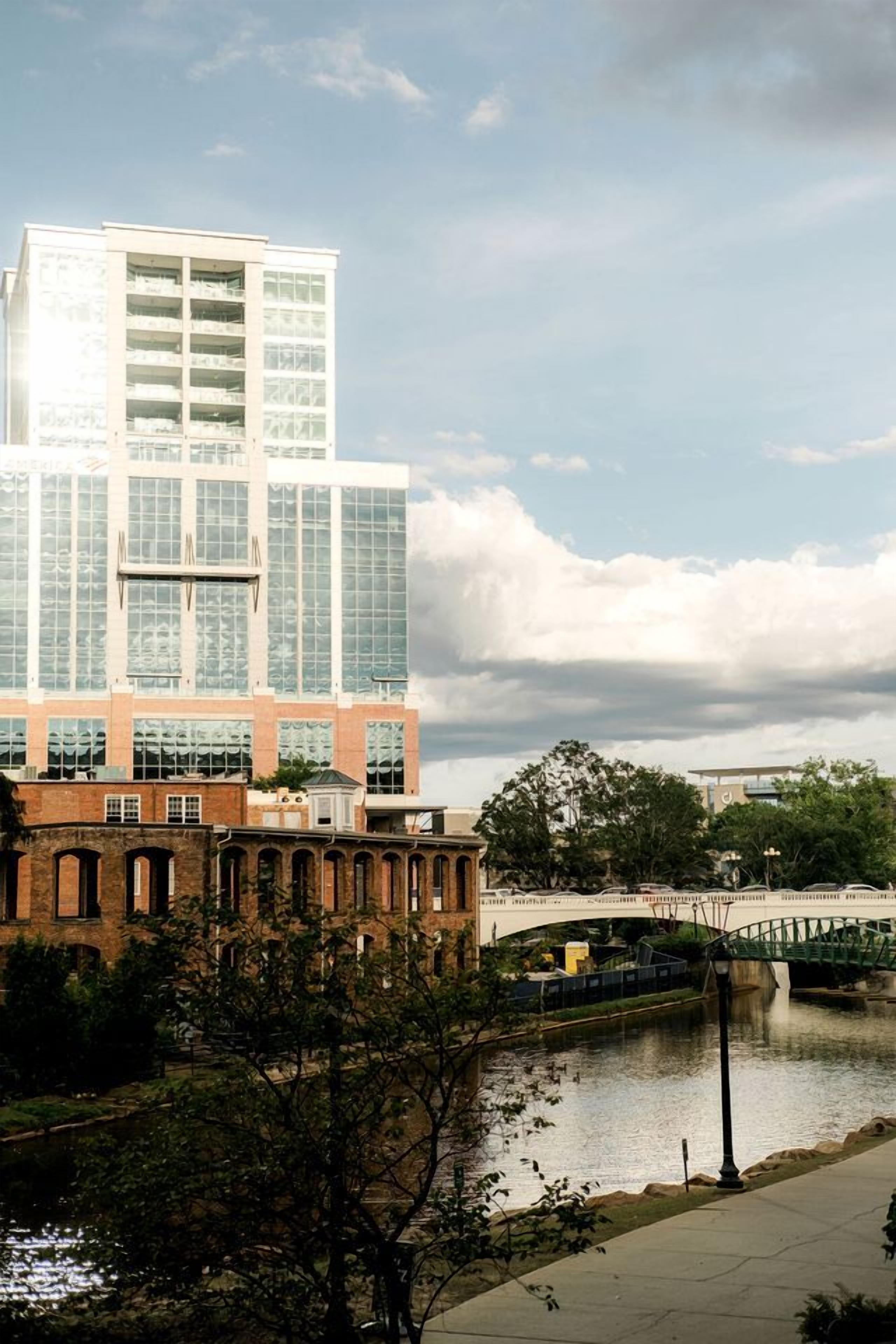 Un edificio moderno con fachada de vidrio, base de ladrillo y un puente sobre una masa de agua.